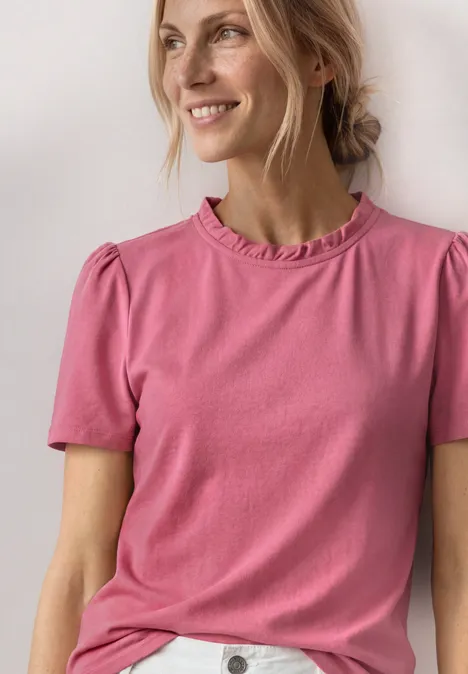 Eine Person trägt ein hellrosa T-Shirt mit kurzen Ärmeln und Rüschen am Rundhalsausschnitt.