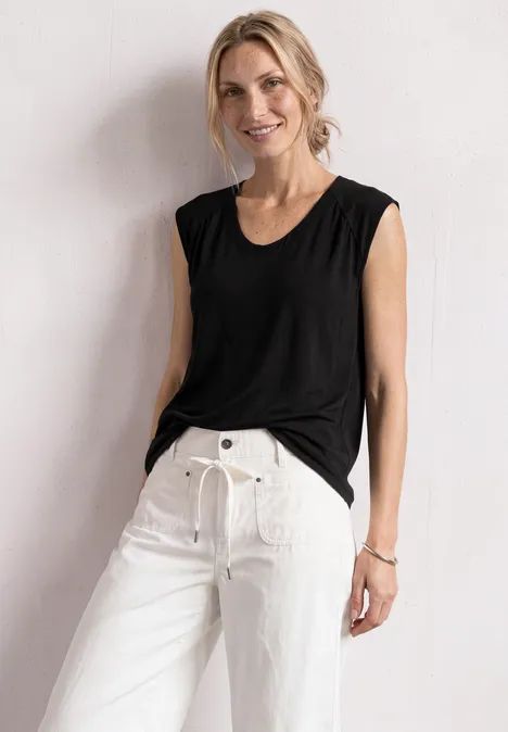 Blonde Frau in schwarzem ärmellosem Top und weißen Hosen lehnt lächelnd an einer Wand.