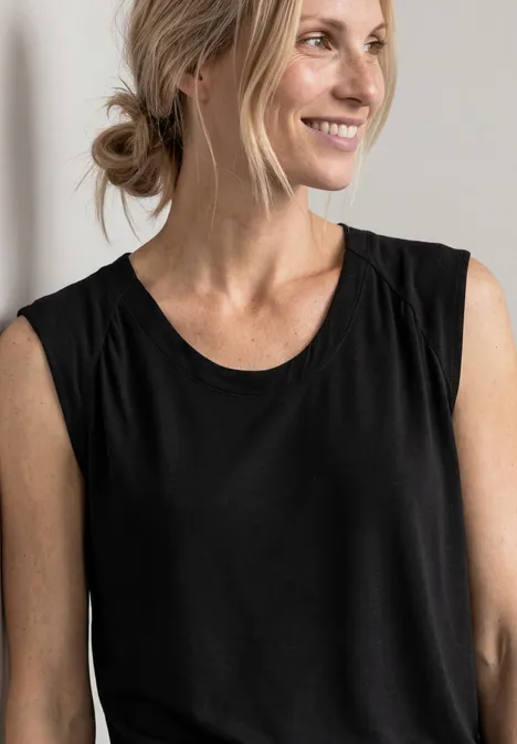 Eine blonde Frau in schwarzem ärmellosem Shirt, lächelnd, vor hellem Hintergrund.