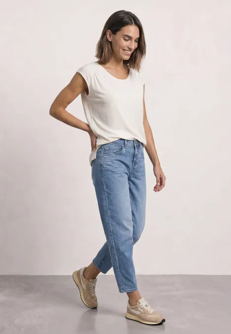 Frau in lockerer Jeans, beigem T-Shirt und Sneakern posiert vor hellem Hintergrund, blickt zur Seite.