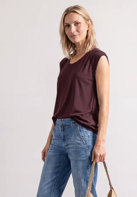 Blonde Frau in Jeans, bordeauxfarbenem Top und Halskette hält eine Handtasche.