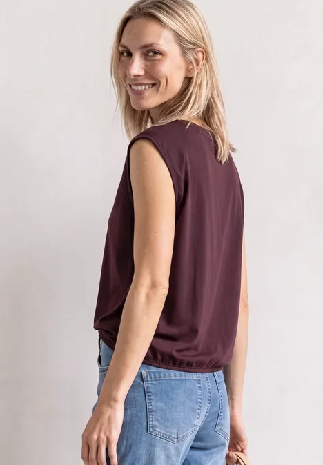 Frau mit blonden Haaren trägt ein ärmelloses Top und Jeans, lächelt über die Schulter.