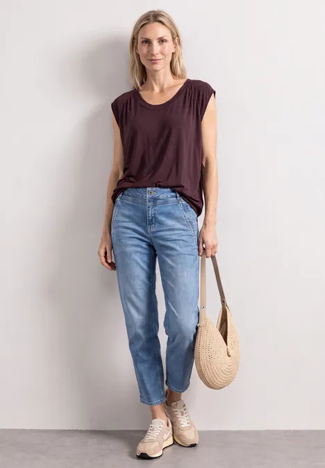 Frau in braunem T-Shirt, hellblauen Jeans, beigen Sneakern und Strohtasche vor hellem Hintergrund.