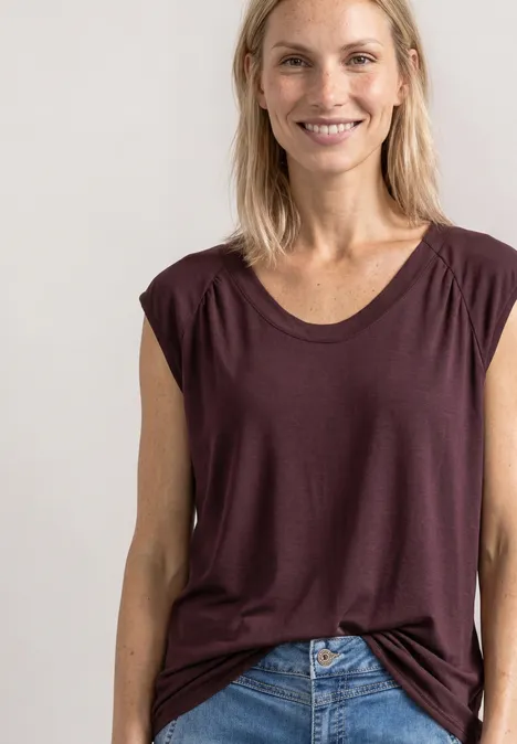 Eine lächelnde Frau im weinroten ärmellosen Top und Jeans vor hellem Hintergrund.