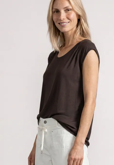 Lächelnde Frau in braunem Top und weißen Hosen vor hellem Hintergrund.