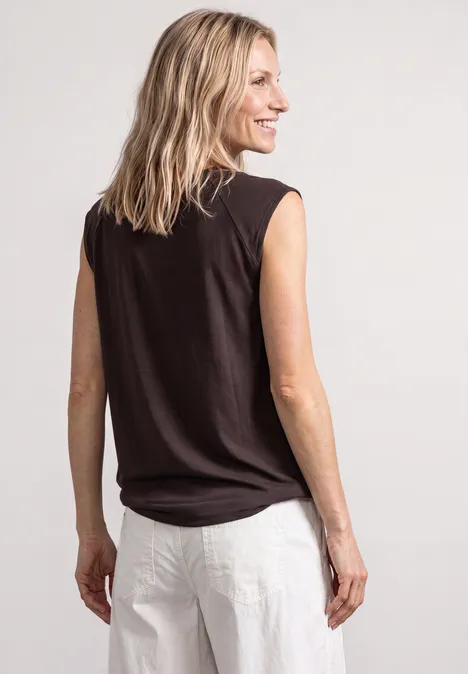 Frau von hinten, lächelnd über die Schulter blickend, in schwarzem ärmellosem Top und weißer Hose.