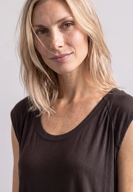 Porträt einer blonden Frau mit Sommersprossen, die in einem dunkelgrauen Top vor hellem Hintergrund leicht lächelt.