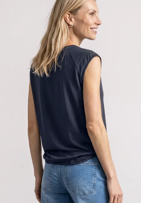 Eine blonde Frau in schwarzem ärmellosem Top und blauen Jeans steht seitlich, lächelt und blickt nach rechts.
