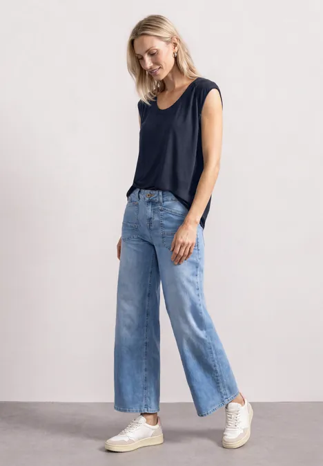 Frau in ärmellosem dunkelblauem Top, weiten Jeans und weißen Sneakers vor neutralem Hintergrund.