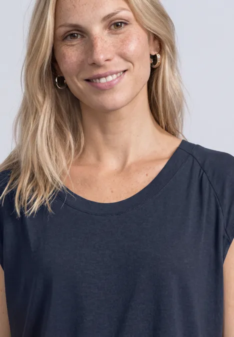 Lächelnde blonde Frau mit Sommersprossen, dunkelblauem Oberteil vor hellgrauem Hintergrund.
