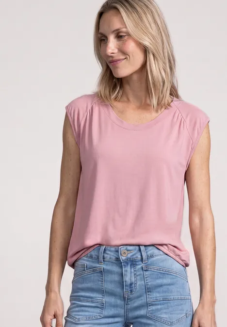Blonde Frau lächelt in ärmellosem, rosafarbenem T-Shirt und blauen Jeans.