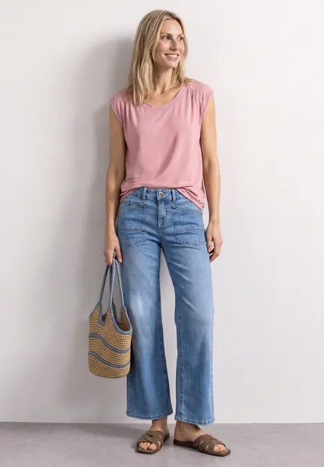 Frau in rosa Top, blauen Schlagjeans und Sandalen trägt eine Strandtasche.