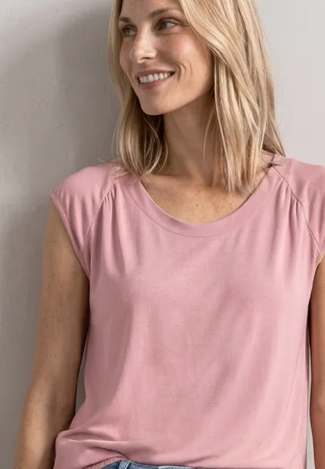 Eine blonde Frau lehnt lächelnd an einer Wand und trägt ein rosafarbenes T-Shirt.