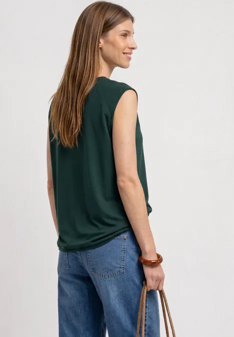 Eine Frau in einem ärmellosen, dunkelgrünen Top und blauen Jeans trägt eine Tasche und blickt seitlich.