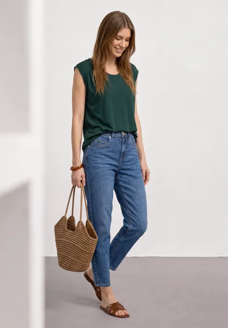 Eine junge Frau in grünem Top und Jeans, mit Strohtasche und Sandalen, steht lächelnd vor neutralem Hintergrund.