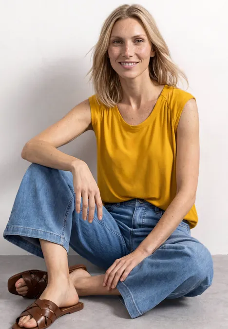 Eine lächelnde Frau sitzt entspannt auf dem Boden, trägt gelbes Top, blaue Jeans und braune Sandalen.