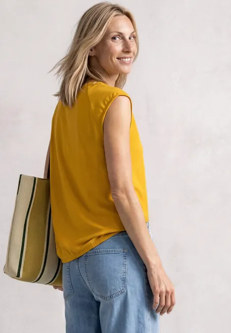 Eine Frau in gelbem Top und Jeans mit gestreifter Tasche schaut lächelnd über die Schulter.