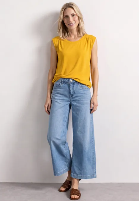 Frau in gelbem ärmellosem Top und weitem Jeans, braune Sandalen, vor neutralem Hintergrund.