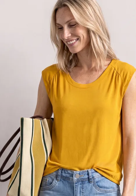 Eine lächelnde blonde Frau in gelbem T-Shirt und Jeans hält eine gestreifte Tasche.
