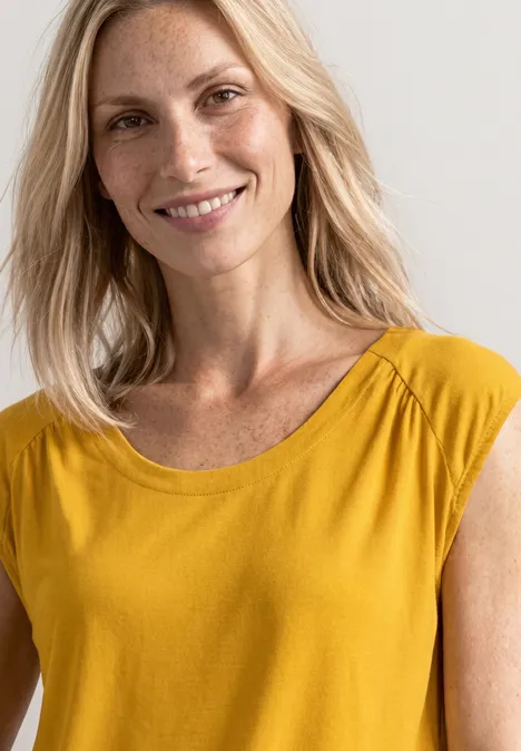 Lächelnde blonde Frau in gelbem Oberteil vor hellem Hintergrund.