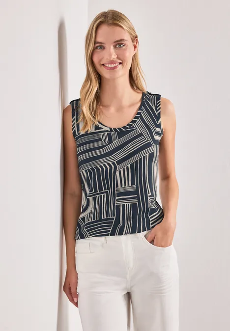 Haut à col en V et boutons décoratifs darkest blue Eine lächelnde Frau in einem ärmellosen, geometrisch gemusterten Top und weißen Hosen lehnt an der Wand.