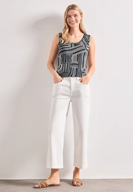 Haut à col en V et boutons décoratifs darkest blue Frau in gemustertem Top, weißen weiten Cropped-Jeans und Sandalen steht mit Händen in den Taschen vor weißer Wand.