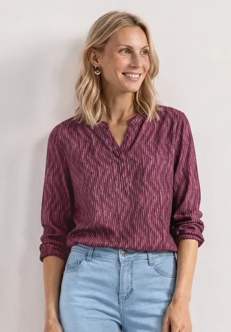 Eine lächelnde Frau mit blondem Haar trägt eine violette gemusterte Bluse und Jeans.