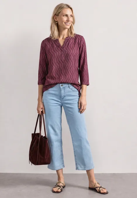 Frau mit purpurroter Bluse, hellblauen Jeans, schwarzen Sandalen und brauner Tasche vor neutralem Hintergrund