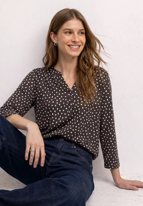 3/4-Arm Shirt mit Split Neck und Print black coffee Eine Frau im gepunkteten Oberteil und Jeans sitzt lächelnd vor weißem Hintergrund.