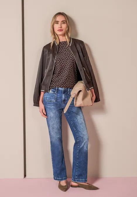 3/4-Arm Shirt mit Split Neck und Print black coffee Frau in gepunkteter Bluse, Jacke überm Schultern, blauen Jeans, beiger Tasche und Ballerinas vor neutralem Hintergrund.