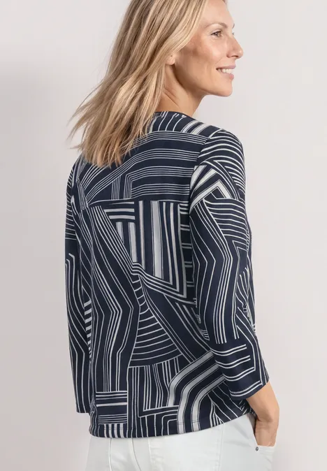 Drei-Viertel-Ansicht einer Frau mit blondem Haar in einer geometrisch gemusterten dunkelblauen Jacke.