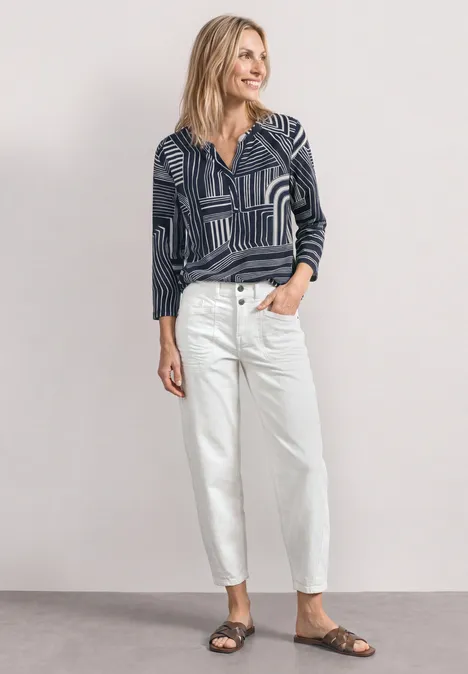 Frau in gemustertem Langarmshirt, weißen Jeans und braunen Sandalen vor neutralem Hintergrund.