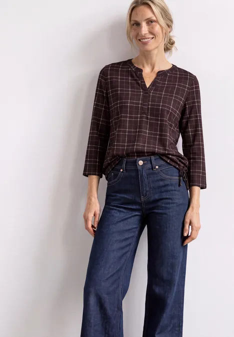 3/4-Arm Shirt mit Split Neck und Print rustic rouge Frau trägt karierte Bluse und blaue Jeans, lächelt vor hellem Hintergrund.