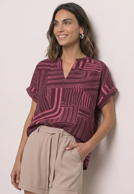 Dropped Shoulder Shirt mit Split Neck blush berry Eine Frau trägt eine gemusterte, rosa-blau gestreifte Bluse mit V-Ausschnitt und sandfarbene Papierbundhose.