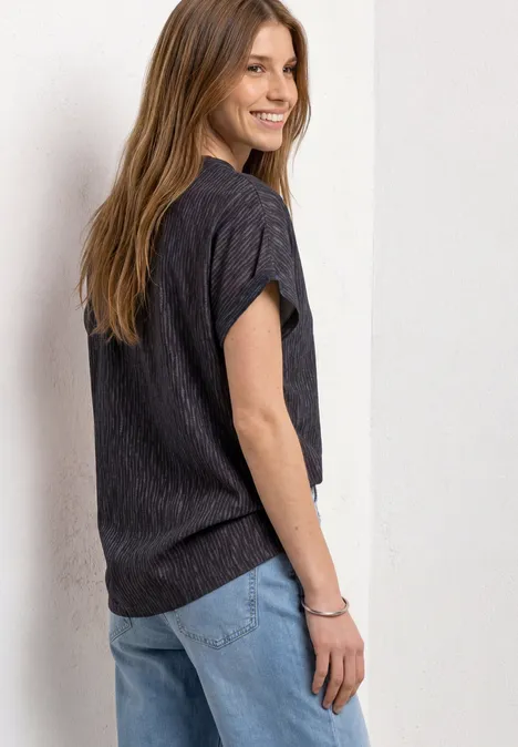Une femme souriante en t-shirt noir et jean s'appuie contre un mur en regardant par-dessus son épaule.