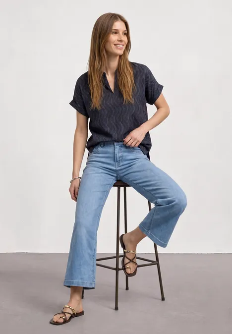 Femme souriante assise sur un tabouret, portant un haut bleu foncé, jean évasé et sandales devant un mur gris clair.