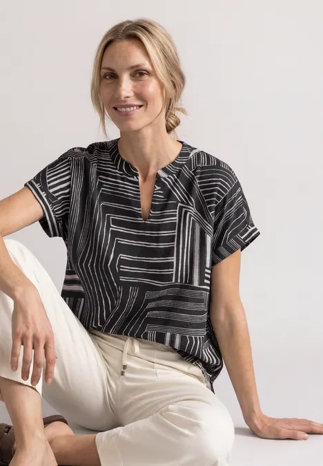 Femme assise, blouse noire à motifs et pantalon clair, sourire détendu.