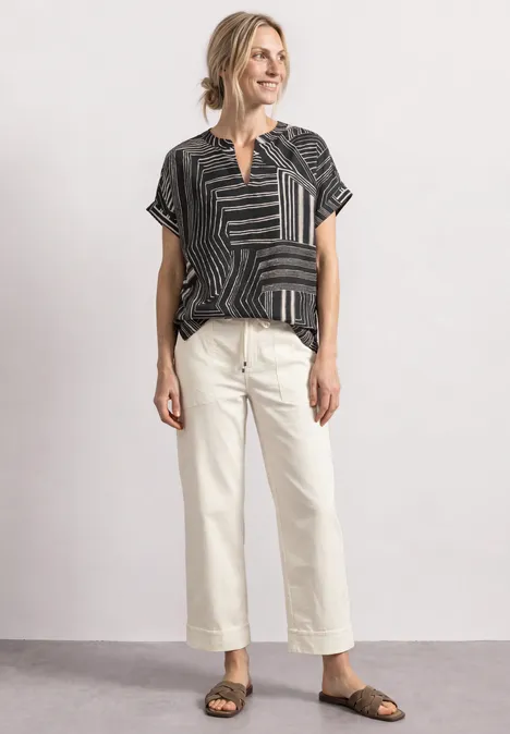 Femme souriante en haut noir à motifs, pantalon court blanc et sandales tressées.