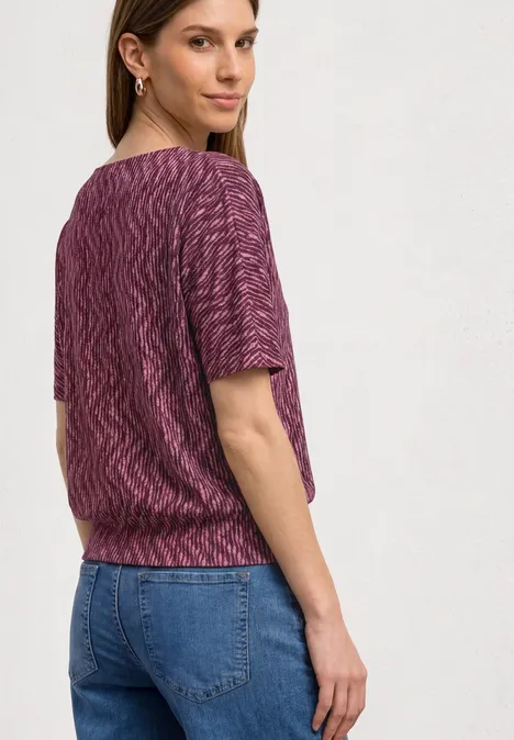 Frau von hinten in lila Strickoberteil und blauen Jeans vor hellem Hintergrund.