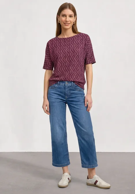 Frau in kurzärmeligem, gemustertem Shirt, weitem Jeans-Crop und weißen Sneakern vor neutralem Hintergrund.
