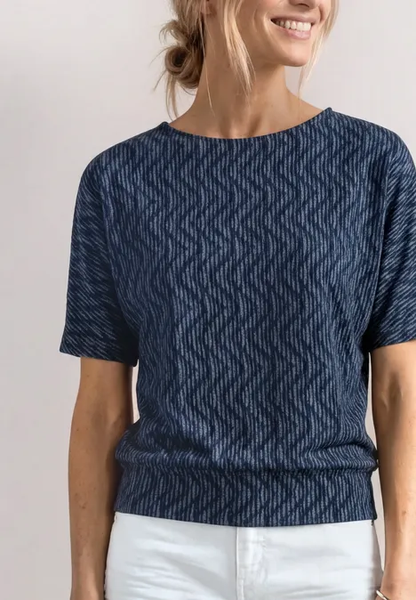 Frau mit lockerer blauer Zickzack-Struktur-Bluse und weißen Hosen, lächelnd und mit zurückgebundenem Haar.