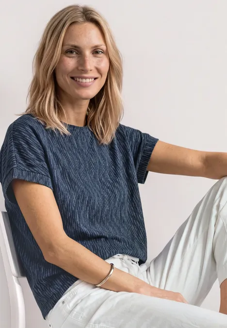 Frau sitzt entspannt auf einem Stuhl, trägt dunkelblaues strukturiertes T-Shirt und weiße Hose.