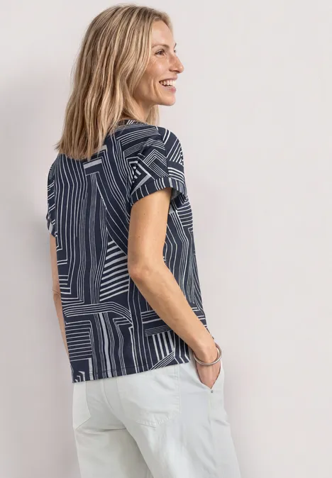 Frau in blau gemustertem T-Shirt und weißen Hosen vor hellem Hintergrund.