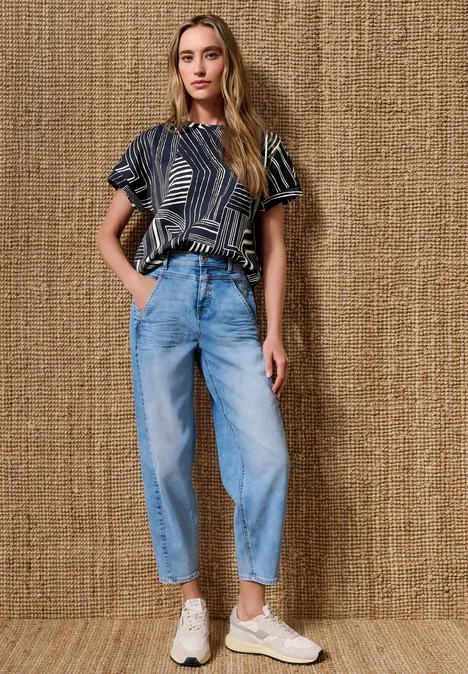 Eine junge Frau trägt eine gemusterte Bluse, weite Jeans und weiße Sneaker vor einem Naturtextil-Hintergrund.