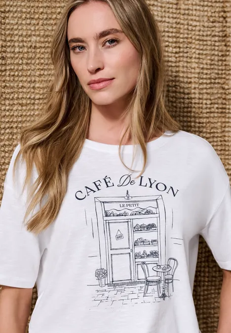 T-Shirt mit Rundhals und Frontprint White Person trägt weißes T-Shirt mit Aufdruck 'Café de Lyon' und Café-Skizze vor geflochtenem Hintergrund