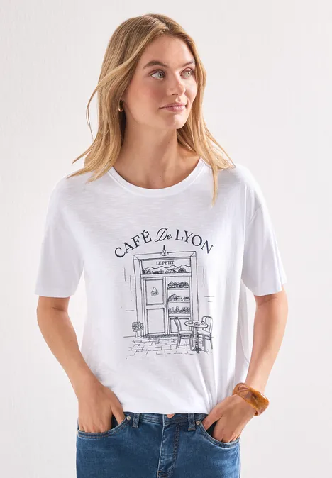 T-Shirt mit Rundhals und Frontprint White Frau in weißem T-Shirt mit Café-de-Lyon-Illustration, Jeanshose und locker fallenden Haaren vor hellem Hintergrund.