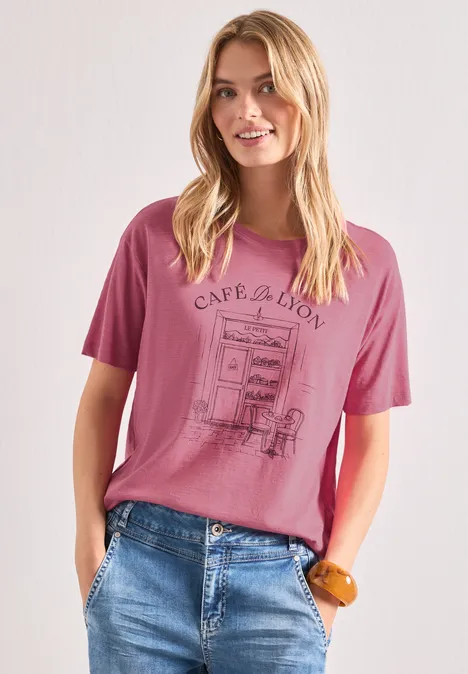 T-Shirt mit Rundhals und Frontprint blush berry Blonde Frau trägt pinkes T-Shirt mit Café-Motiv und blaue Jeans, mit Händen in den Taschen.