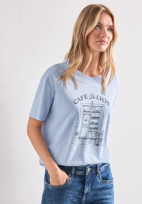 T-Shirt mit Rundhals und Frontprint whispery blue Frau in hellblauem T-Shirt mit 'CAPE DE LYON'-Aufdruck und Jeans vor weißer Wand