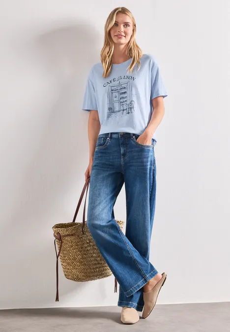 T-Shirt mit Rundhals und Frontprint whispery blue Frau mit unkenntlichem Gesicht trägt hellblaues T-Shirt, weite Jeans, Basttasche und Slipper.