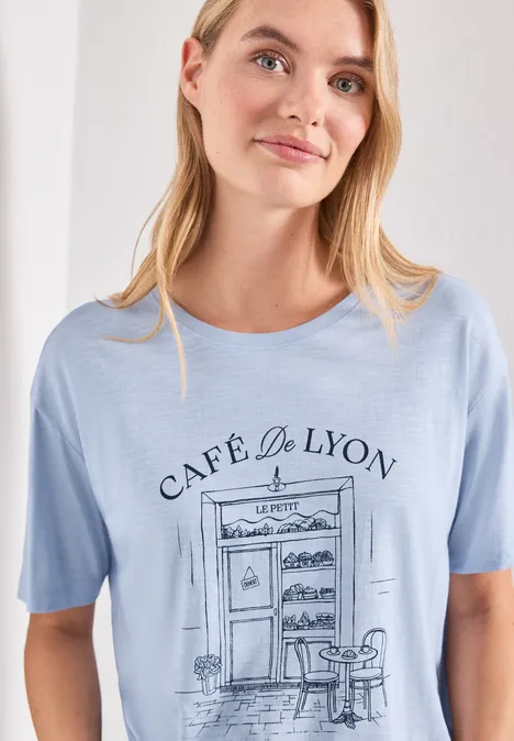 T-Shirt mit Rundhals und Frontprint whispery blue Frau trägt hellblaues T-Shirt mit Café-Skizze 'Café de Lyon', inklusive Tisch und Stuhl.